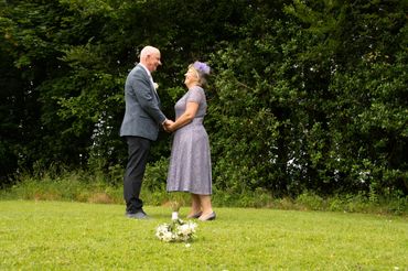 Couple holding hands and smiling outdoors with bouquet on grass.