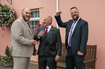 Three men in suits posing happily outside a pink building.