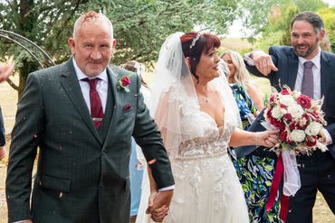 Newlyweds holding hands amidst wedding celebrations with guests throwing confetti.