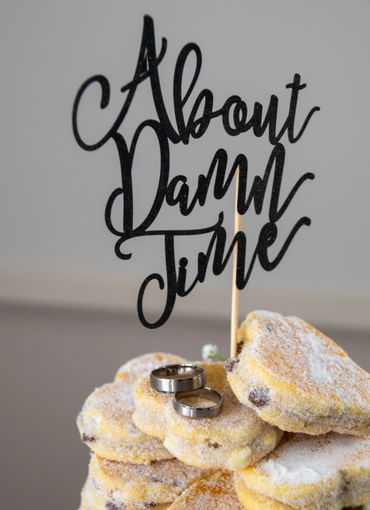 Wedding rings on powdered sugar-dusted pastries with 'About Damn Time' cake topper.