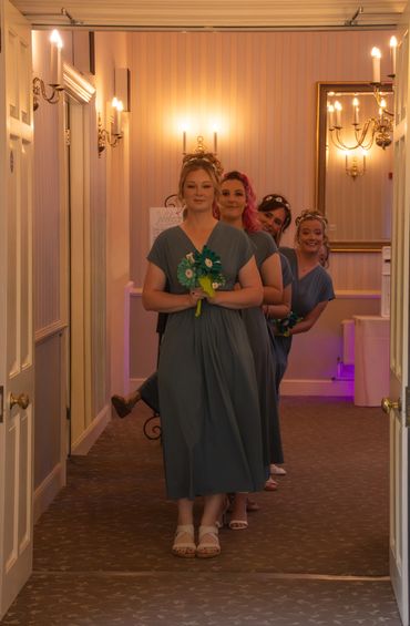 Four bridesmaids in blue dresses lined up holding bouquets in a warmly lit hallway.