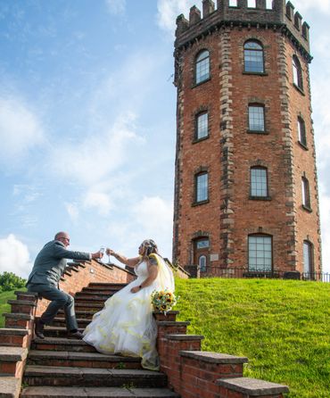 Bride and groom toast on steps near a historic brick tower on a sunny day.