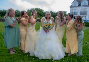 Bride in white gown surrounded by bridesmaids blowing kisses outdoors.