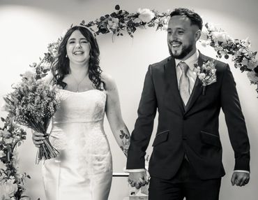 A joyful bride and groom holding hands under a floral arch at their wedding.