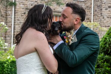 Bride and groom lovingly kiss their small black dog during wedding.