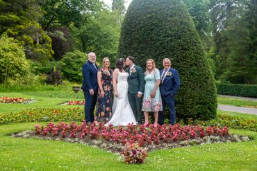 Newlyweds kiss surrounded by family in a lush garden with vibrant flowers.