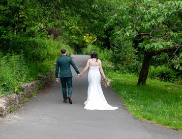 Newlyweds walking hand in hand down a tree-lined path.