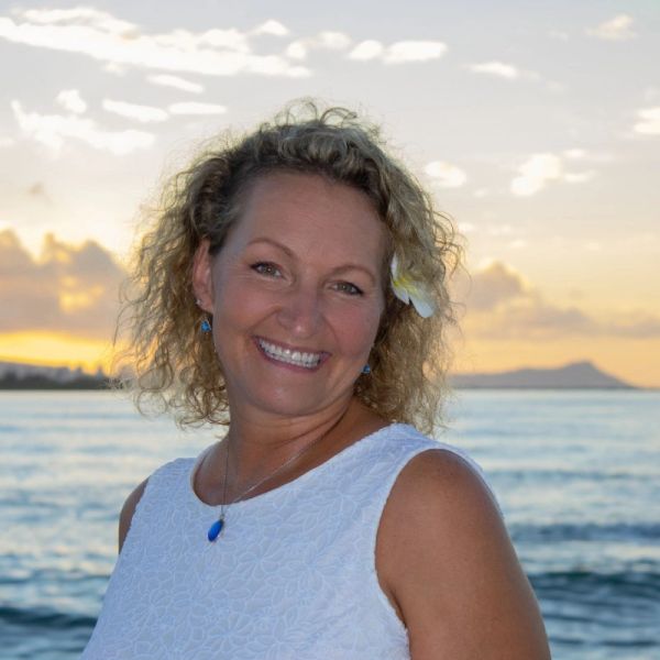Smiling woman with curly hair and flower in hair at sunset by the ocean.