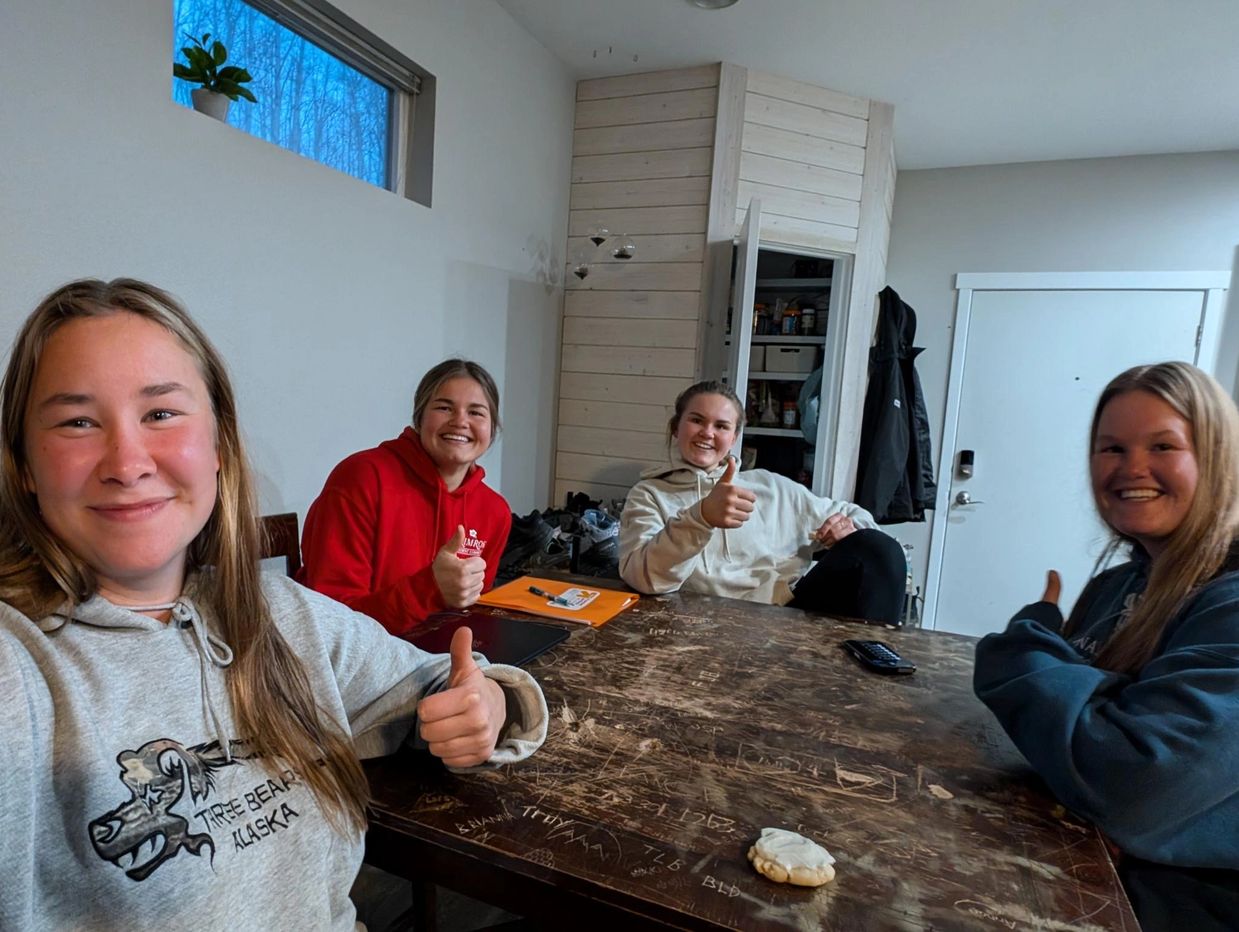 Four smiling women sitting around a table giving thumbs up inside a cozy room.