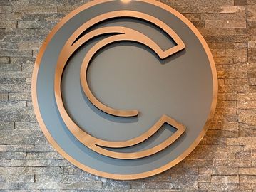 A wooden logo of overlapping letter C on a gray circular background.