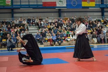 pokaż aikido na hali sportowej, setka ludzi na widowni: rzuca młoda kobieta, pada człowiek w hakamie