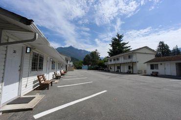 Empty motel parking lot with white buildings and mountain backdrop under a partly cloudy sky.