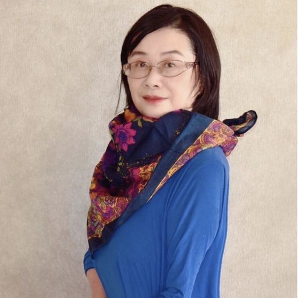 A woman wearing glasses, a blue top, and a colorful scarf poses against a neutral background.
