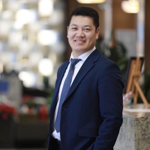 Smiling man in a navy blue suit standing indoors with hands in pockets.