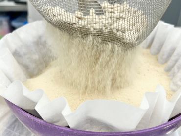 Flour being sifted through a metal strainer into a paper-lined purple bowl.