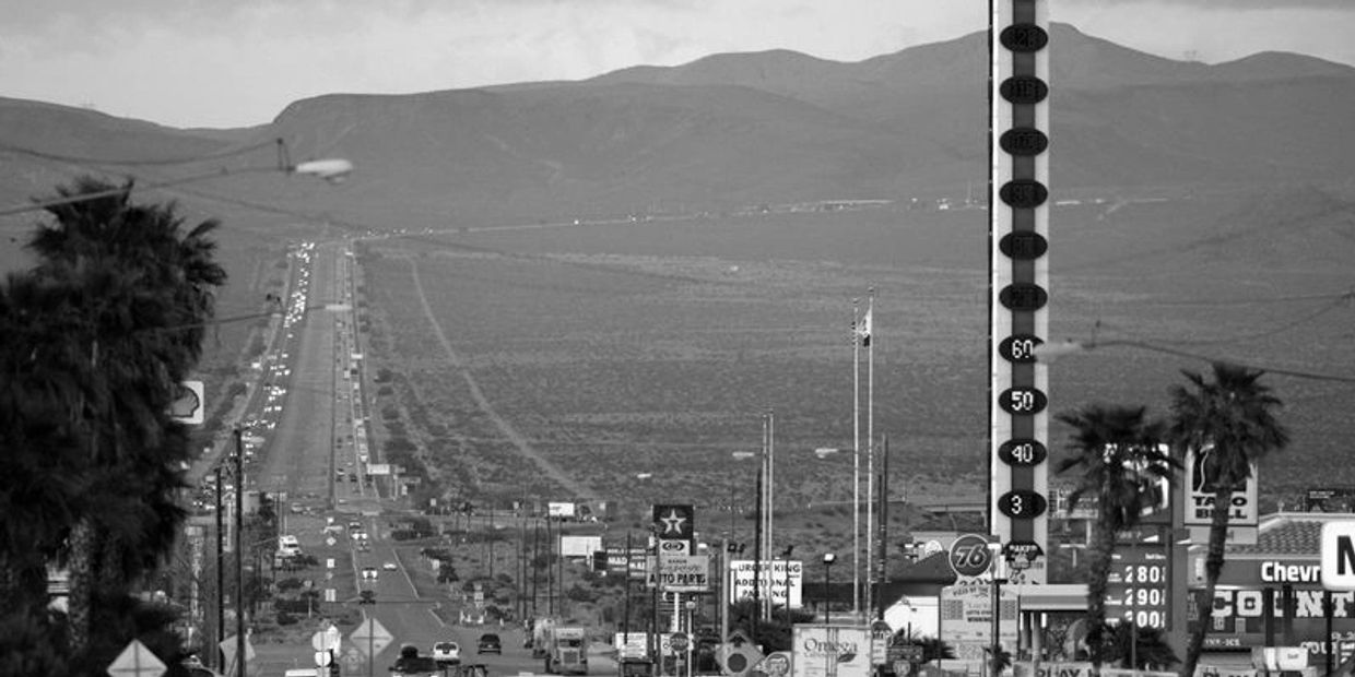 Long highway stretching through a desert town with gas station signs and mountains in the background.