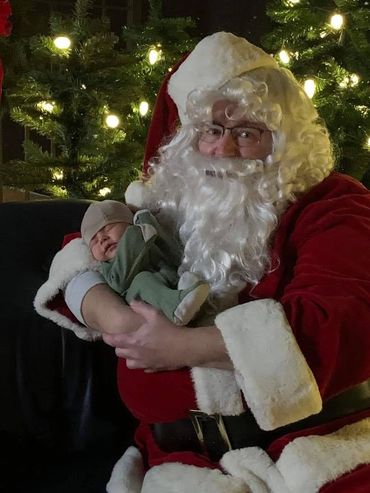 Santa Claus holds a peacefully sleeping baby in front of a decorated Christmas tree.