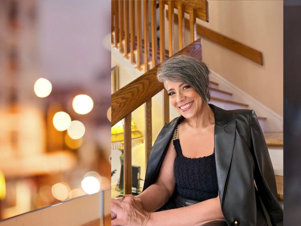 Smiling woman with short gray hair sitting by a wooden staircase indoors.