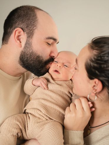 Parents tenderly holding and kissing their newborn baby in matching beige outfits.