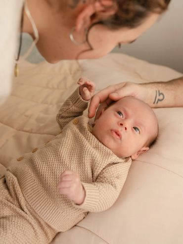 A baby lying on a bed looking at an adult with a hand gently touching their head.