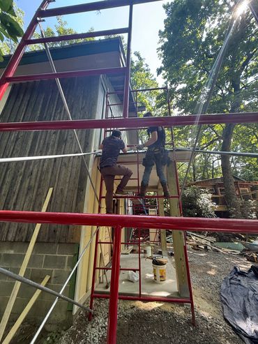 Two workers on red scaffolding working on a house exterior under sunlight.