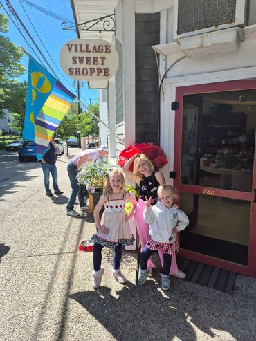 Three happy children posing outside Village Sweet Shoppe on a sunny day.