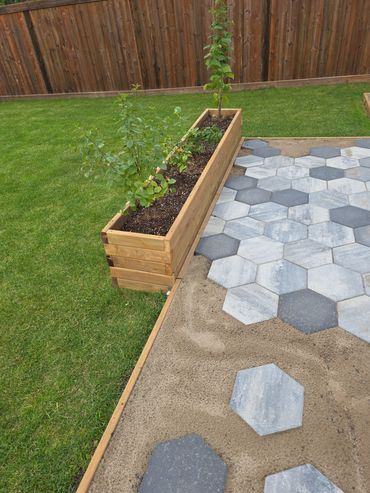 Hexagonal stone tiles being laid next to a wooden planter box in a garden.