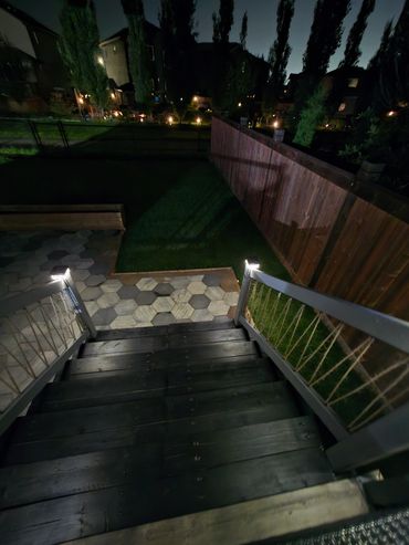 Night view down wooden stairs to lit backyard patio with hexagonal tiles, custom rope design in plac