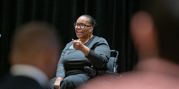 Dr. Pearson speaking on stage in wheelchair. Blurred images of audience members in foreground.