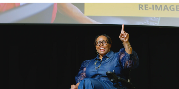Dr. Pearson speaking on stage in wheelchair. Re-IMAGE slide is on large screen in background.