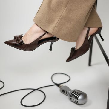 Brown high-heeled shoes with a bow detail worn with beige pants, next to a vintage microphone on the floor.