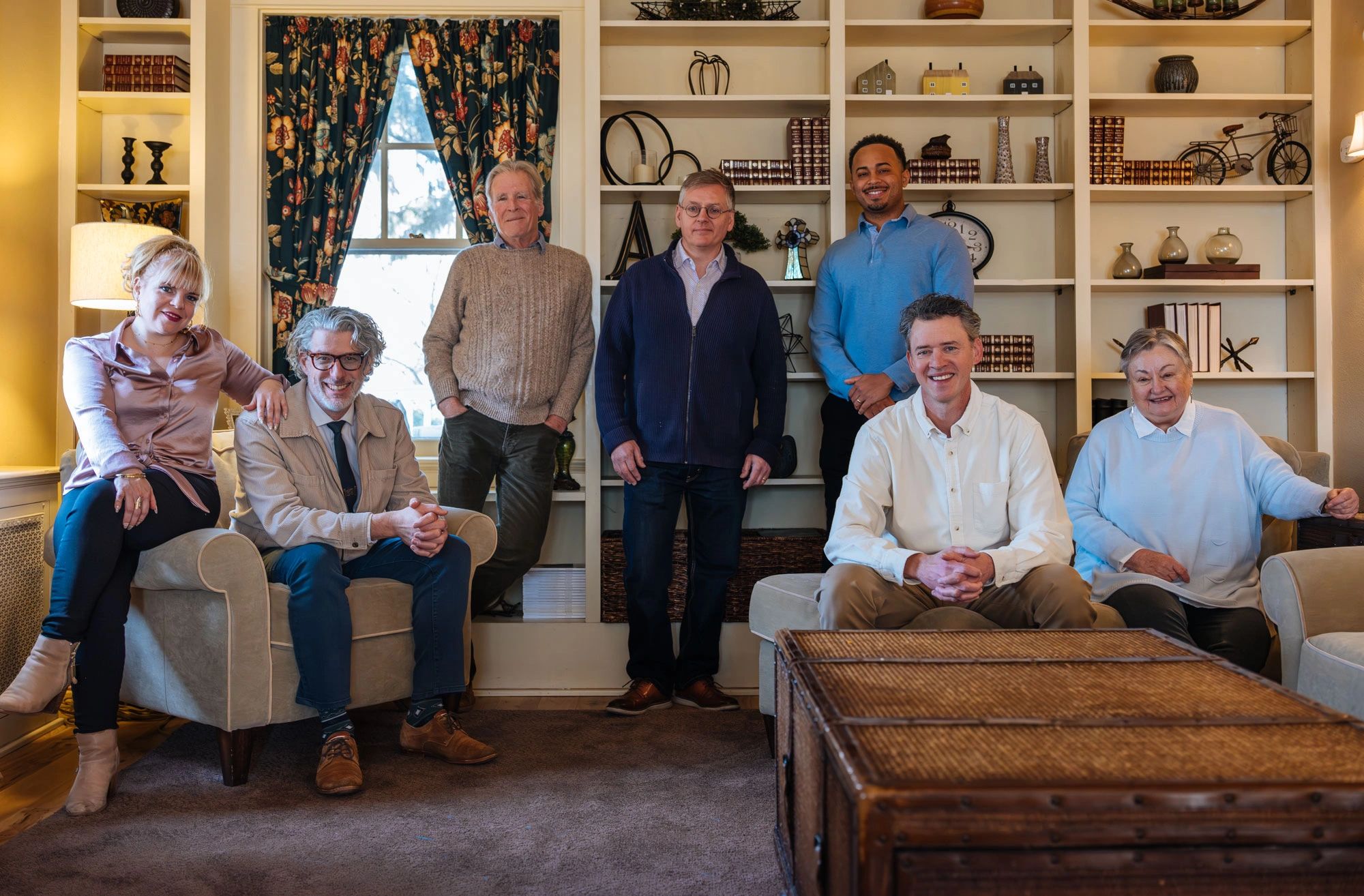 Group of seven people posing in a cozy living room with bookshelves and floral curtains.