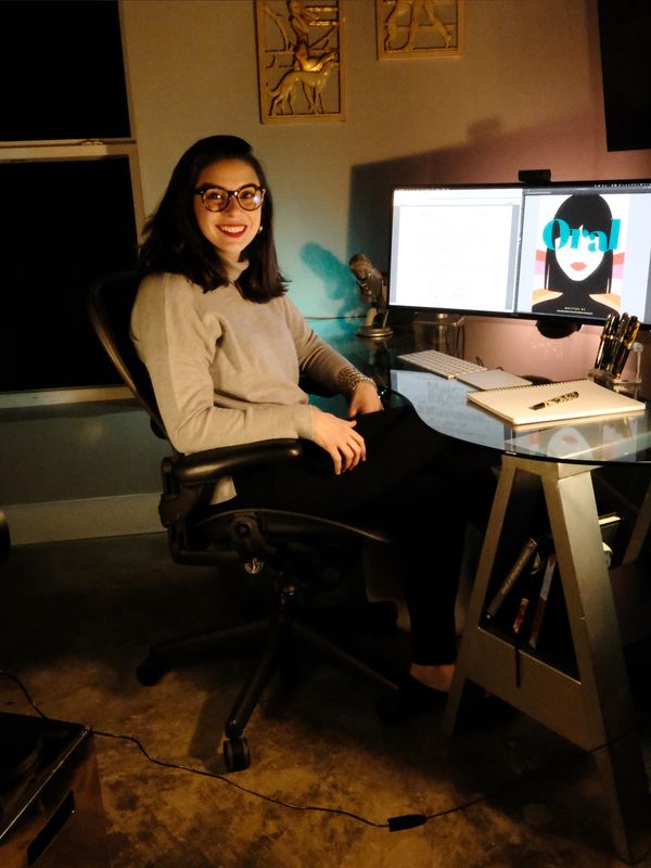 A dark-haired woman wearing glasses sits in front of her computer.