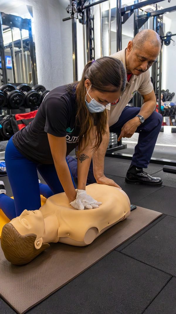 A woman practices CPR on a training dummy under instructor supervision.