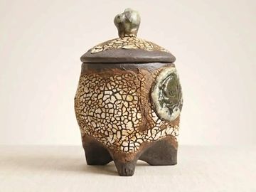 A rustic, textured ceramic jar with a lid and three legs.
