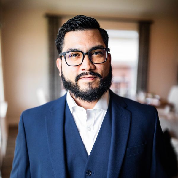 Man with glasses and beard wearing a navy blue suit indoors.