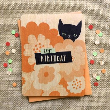 A wooden happy Birthday card featuring a black cat and flowers.