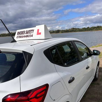 White driving school car by a lakeside under cloudy sky.