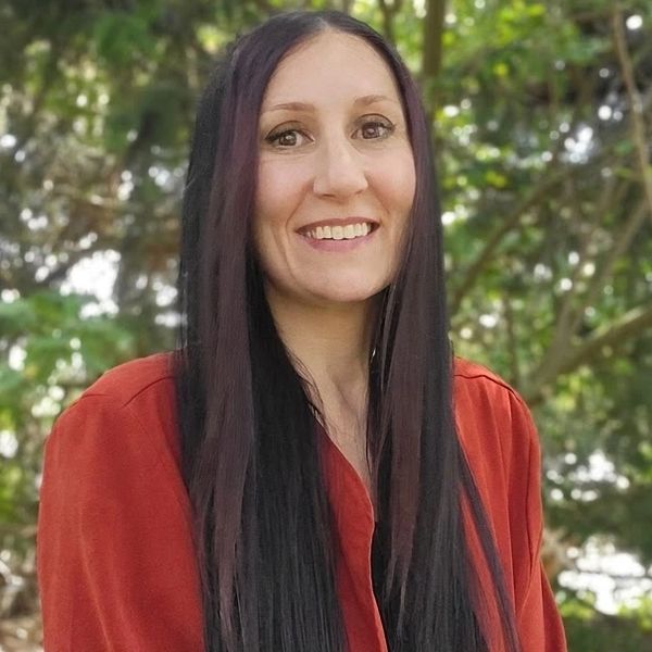 Smiling woman with long dark hair in an orange blouse outdoors.