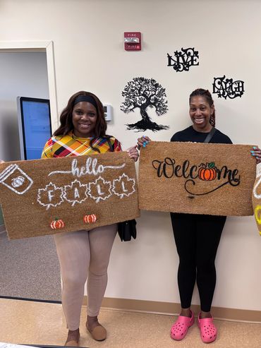 Three women holding decorated fall-themed doormats indoors.