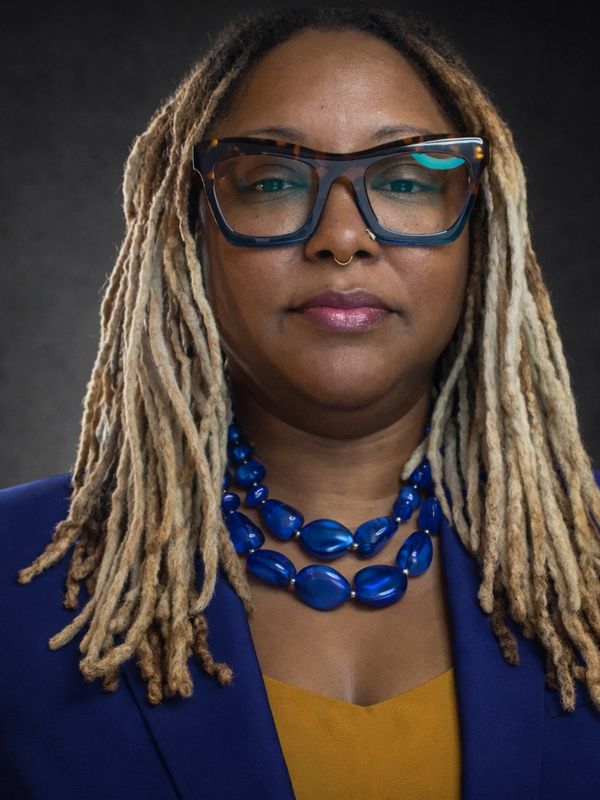 Confident woman with blonde dreadlocks, blue glasses, and bold blue necklace.