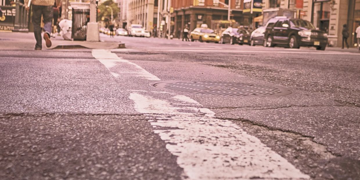 Close-up of a city street with worn white crosswalk lines and cars in the background.