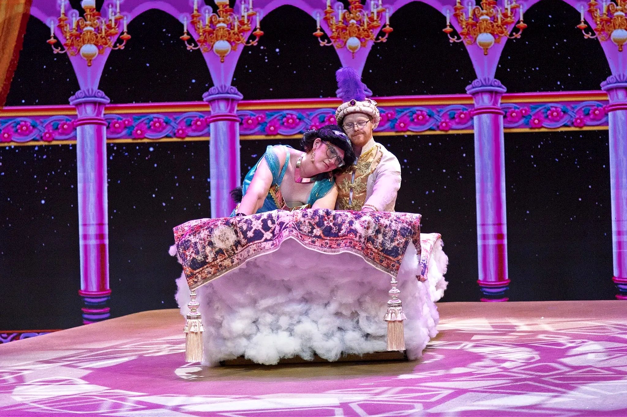 A man and a woman in Aladdin and Jasmine costumes ride a magic carpet over a cloud on a stage.