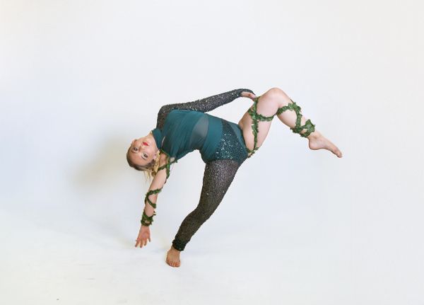 A woman in a green and black outfit performs a balancing dance pose with vines wrapped around her limbs.