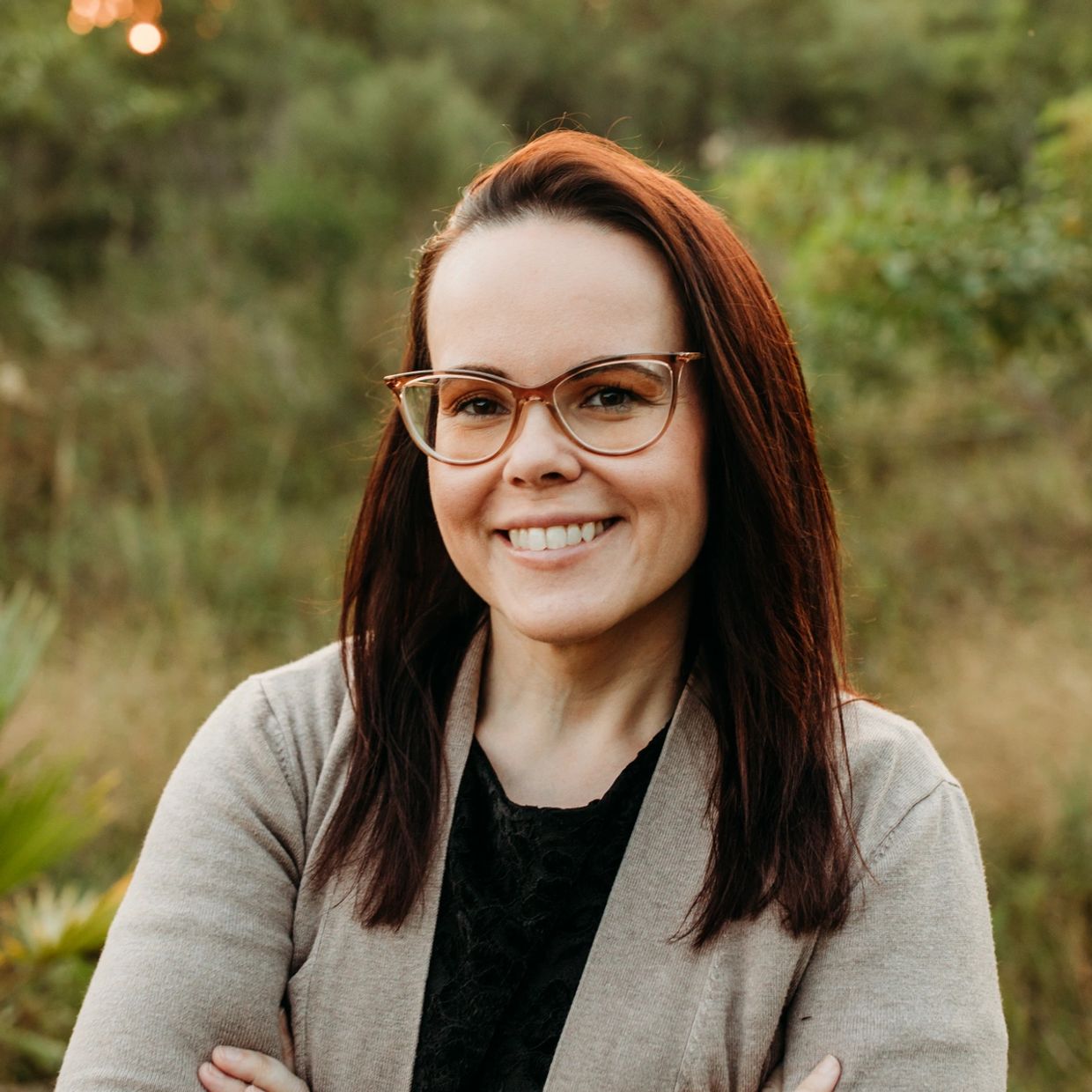Smiling woman with glasses standing outdoors with arms crossed.