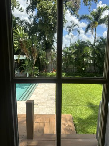 View through a window of a lush backyard with a pool and palm trees.