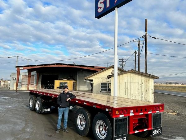Man standing next to a flatbed trailer outside industrial buildings.