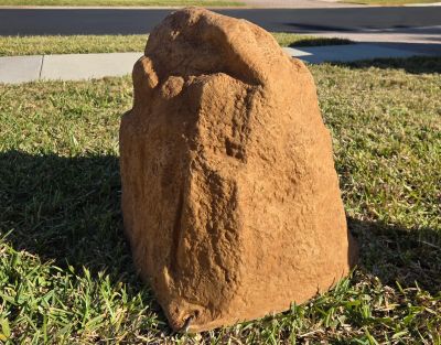 Large irregular rock on grass near a sidewalk and street.