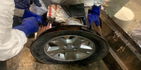 Two individuals in protective gear handling a car tire with a suspicious package inside.