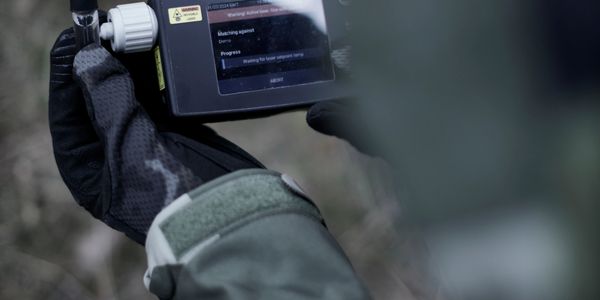 Person in camouflage holding a handheld device with a warning label.